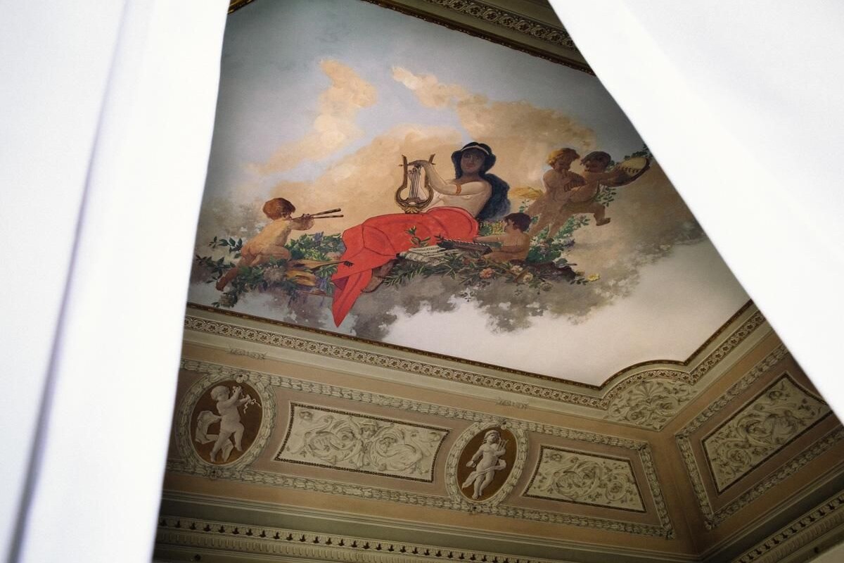 A ceiling with a fresco of a woman playing a lyre, surrounded by cherubs and foliage.