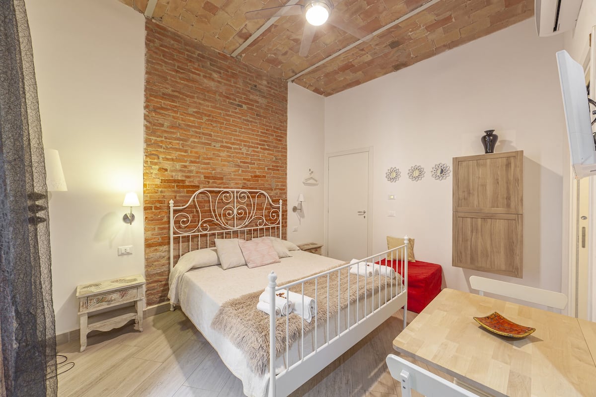 A bedroom with a brick wall, a bed with a white metal frame, a wooden cabinet, and a small table with a bowl.