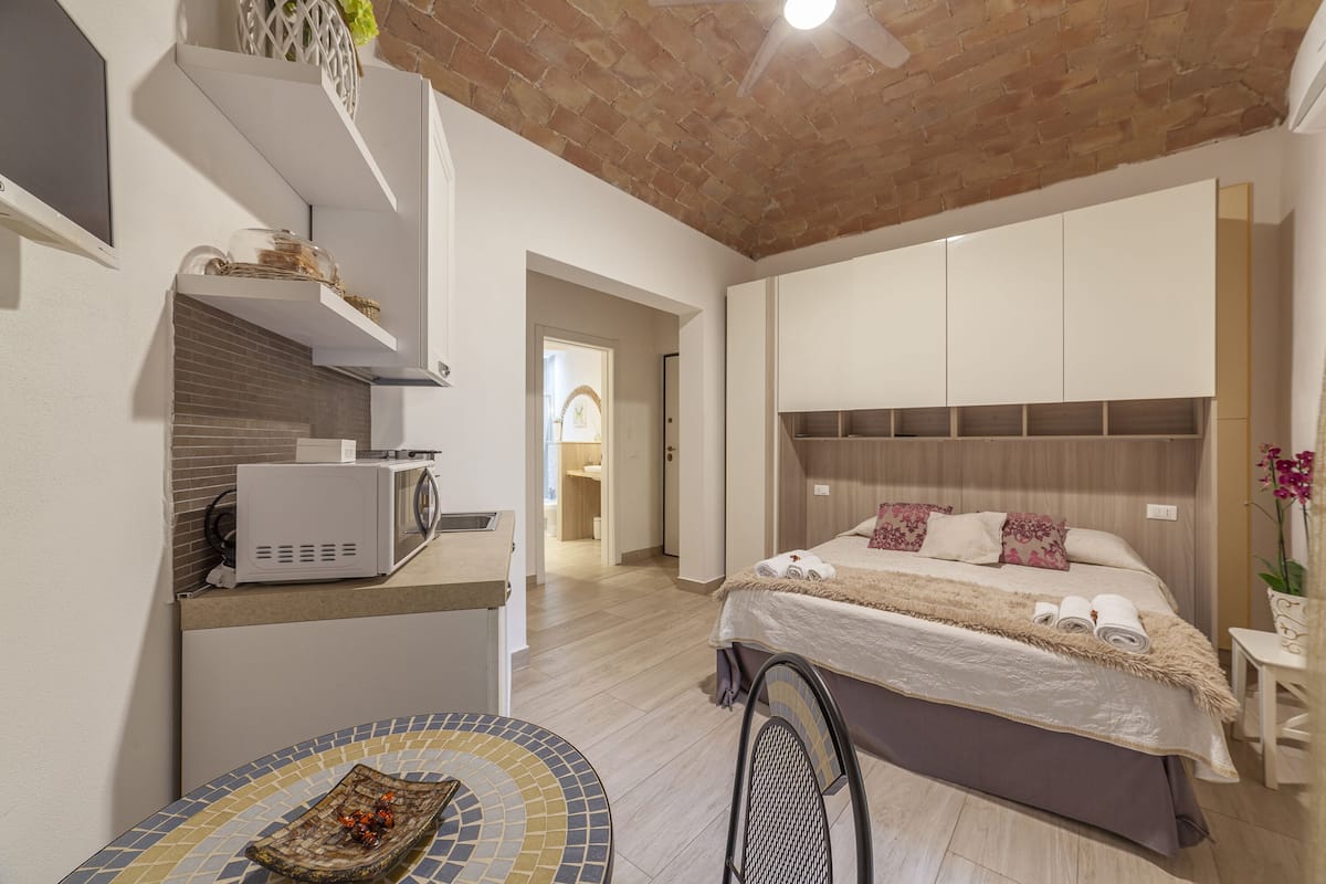 A modern hotel room with a kitchen area, a bed with a large headboard, and a small dining table.