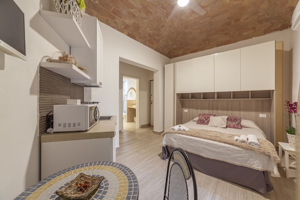 A modern hotel room with a kitchen area, a bed with a large headboard, and a small dining table.