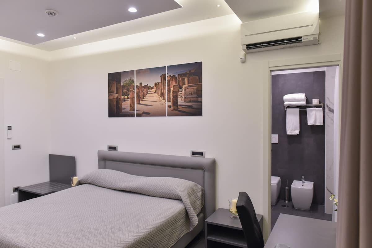 A modern hotel room with a large bed, a bedside table, a small desk, and a bathroom with a shower, toilet, and sink.