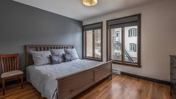 Un dormitorio con una cama de madera, una silla y ventanas amplias que dan a vistas de edificios.