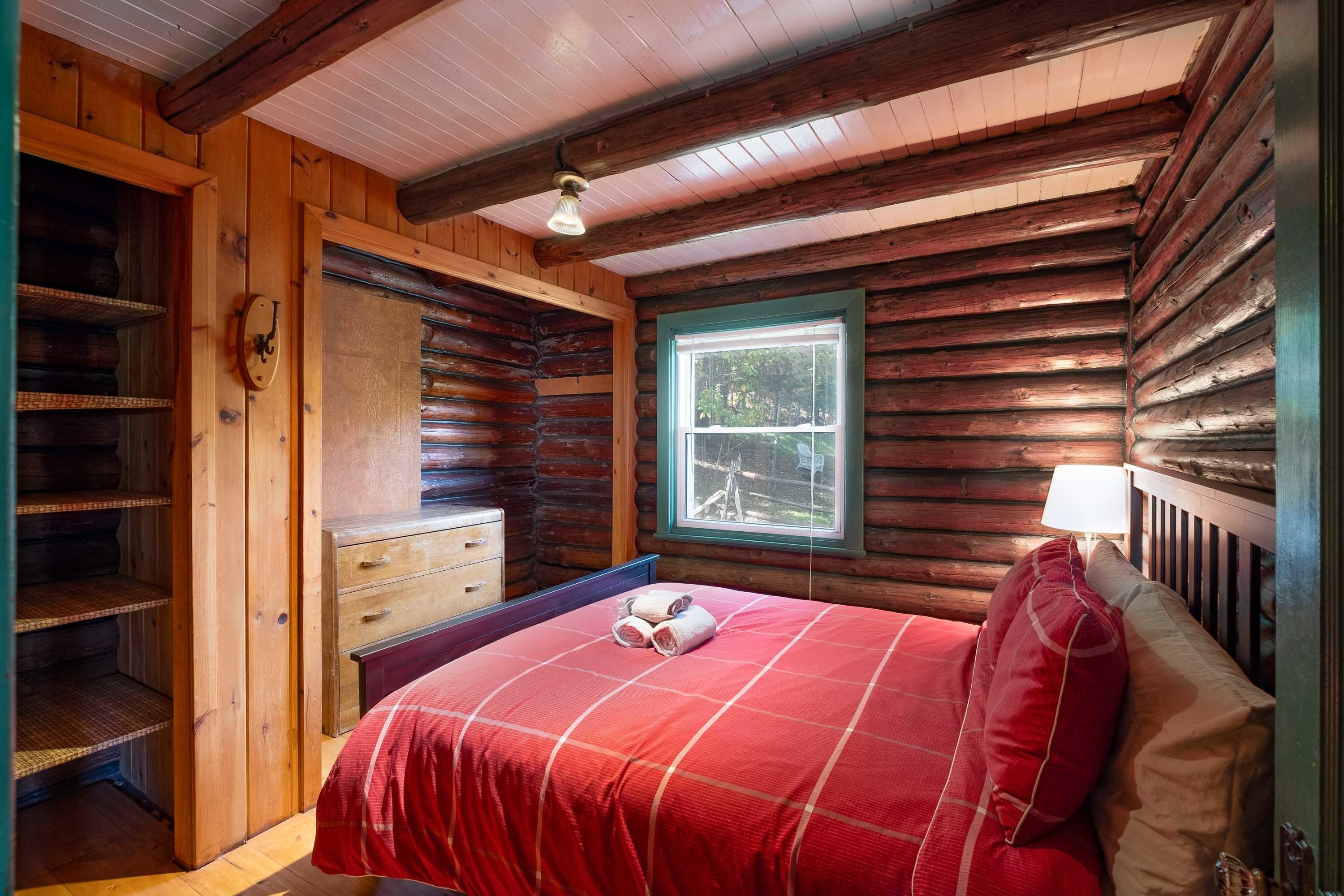 A log cabin bedroom with a bed, a dresser, a window, and a lamp.