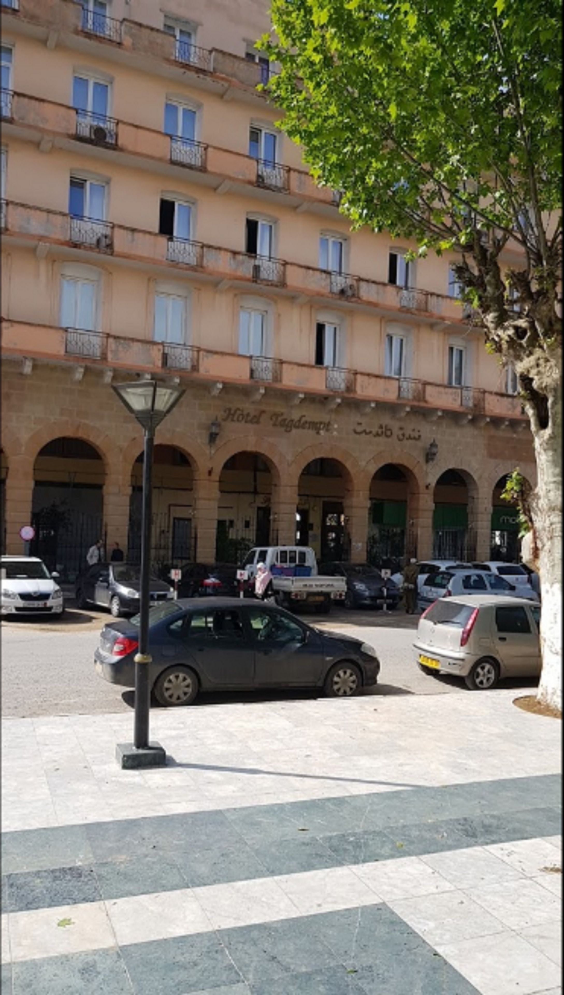 A multi-story hotel with arched windows and balconies, a parking area with several cars, and a pedestrian crossing.