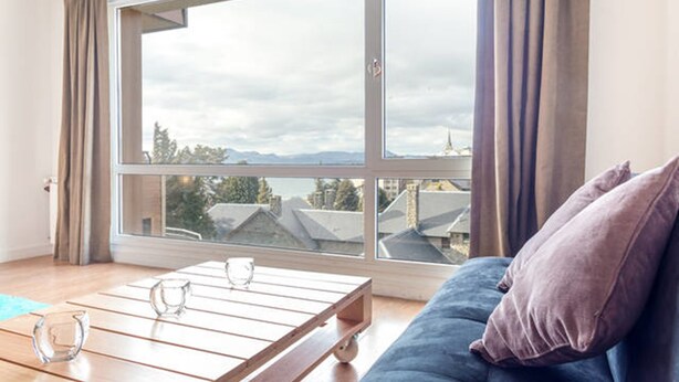 A room with a large window, a blue sofa, and a wooden coffee table with two glasses. - Estudio A 20M Del Centro Cívico (San Carlos de Bariloche)