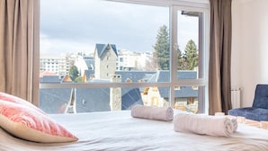 A hotel room with a large window, a bed with pillows, a chair, and a view of buildings and trees. - Estudio A 20M Del Centro Cívico (San Carlos de Bariloche)