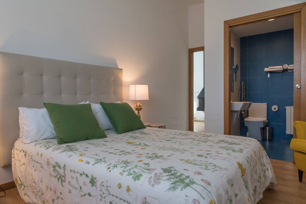 Une chambre à coucher avec un lit, des oreillers, une lampe de chevet, une porte en bois et une salle de bain visible à travers une porte ouverte.
