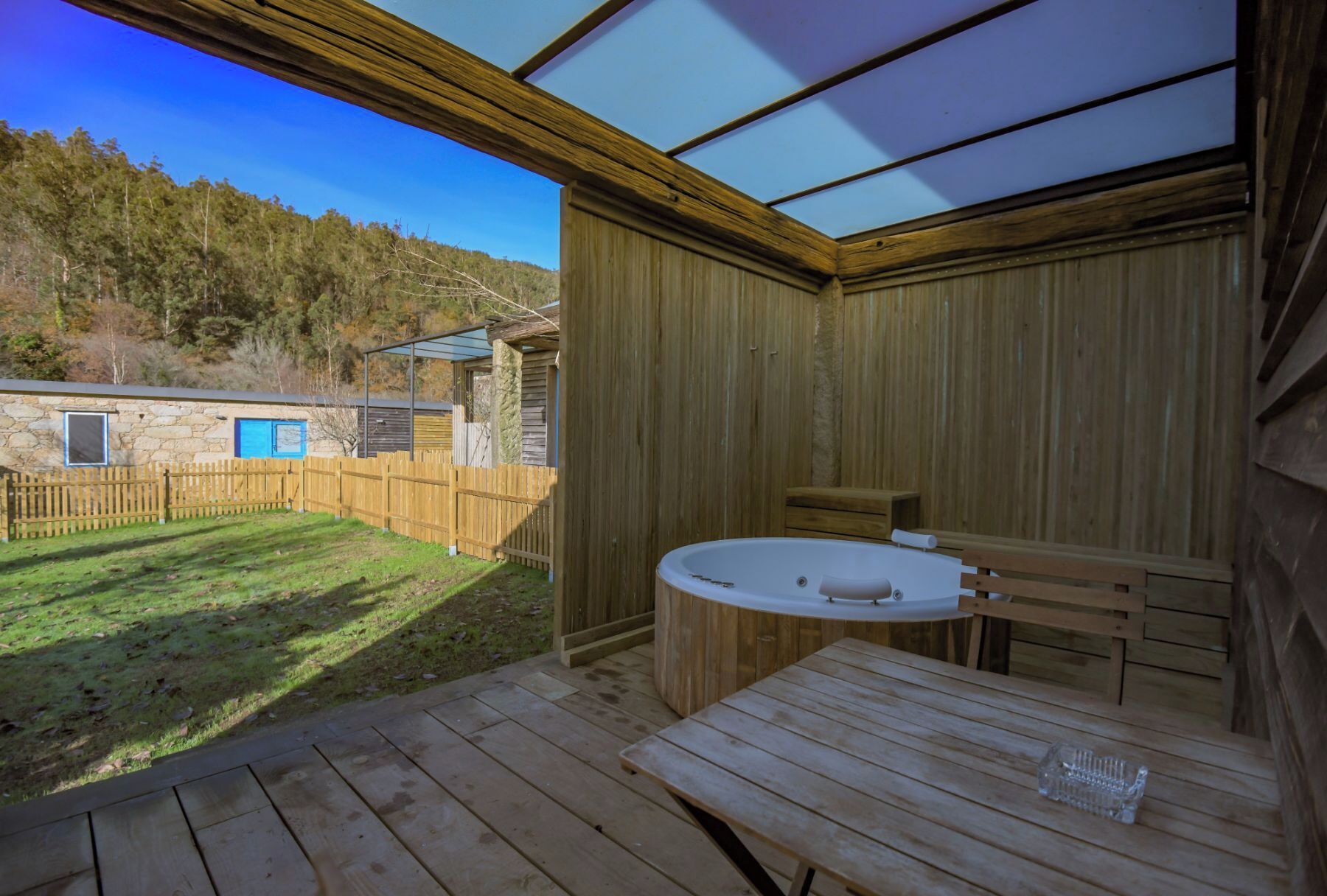A wooden cabin with a hot tub and a table, surrounded by a wooden deck and a fenced yard.