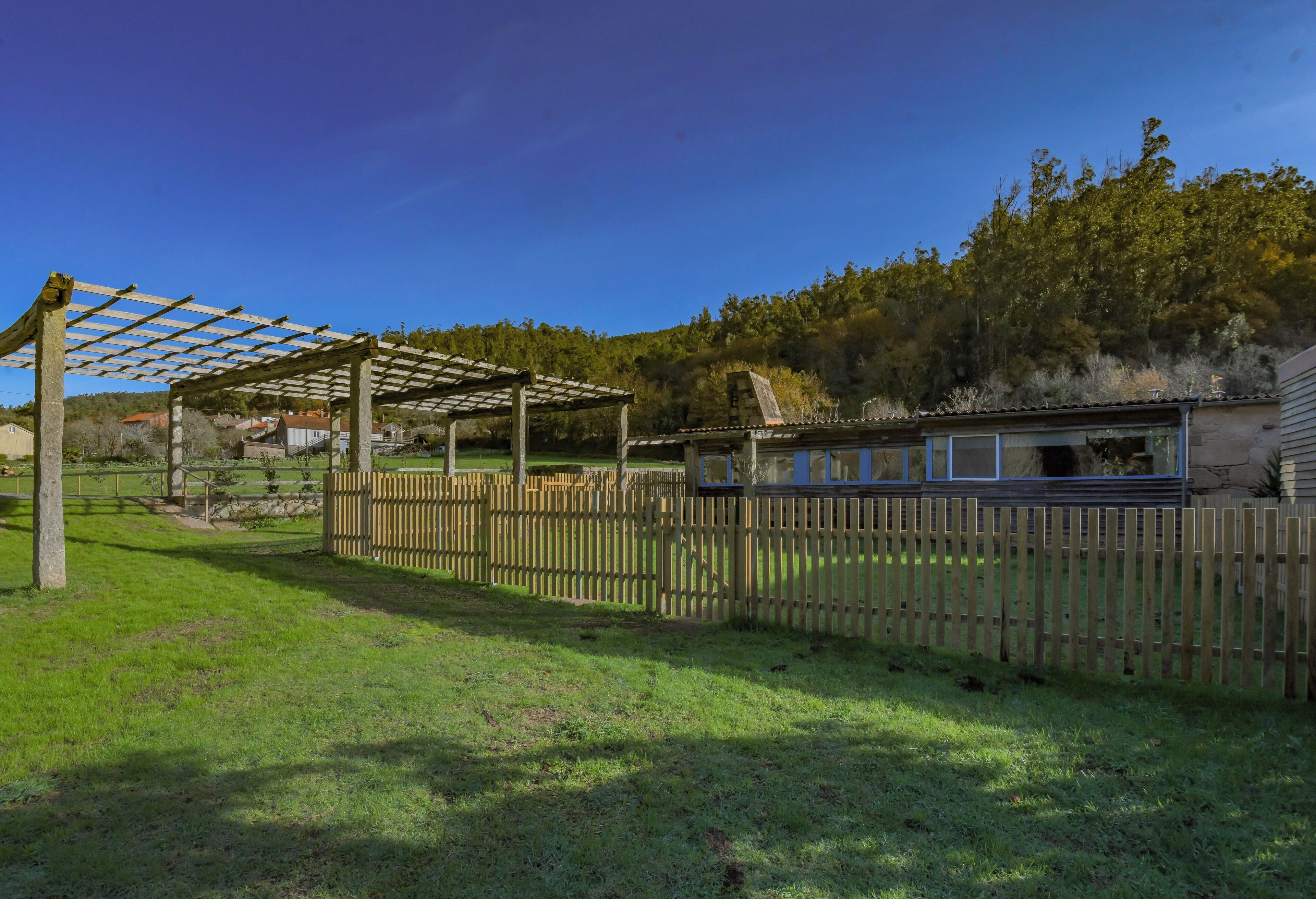 A wooden pergola structure in a grassy area with a wooden fence and a building in the background.