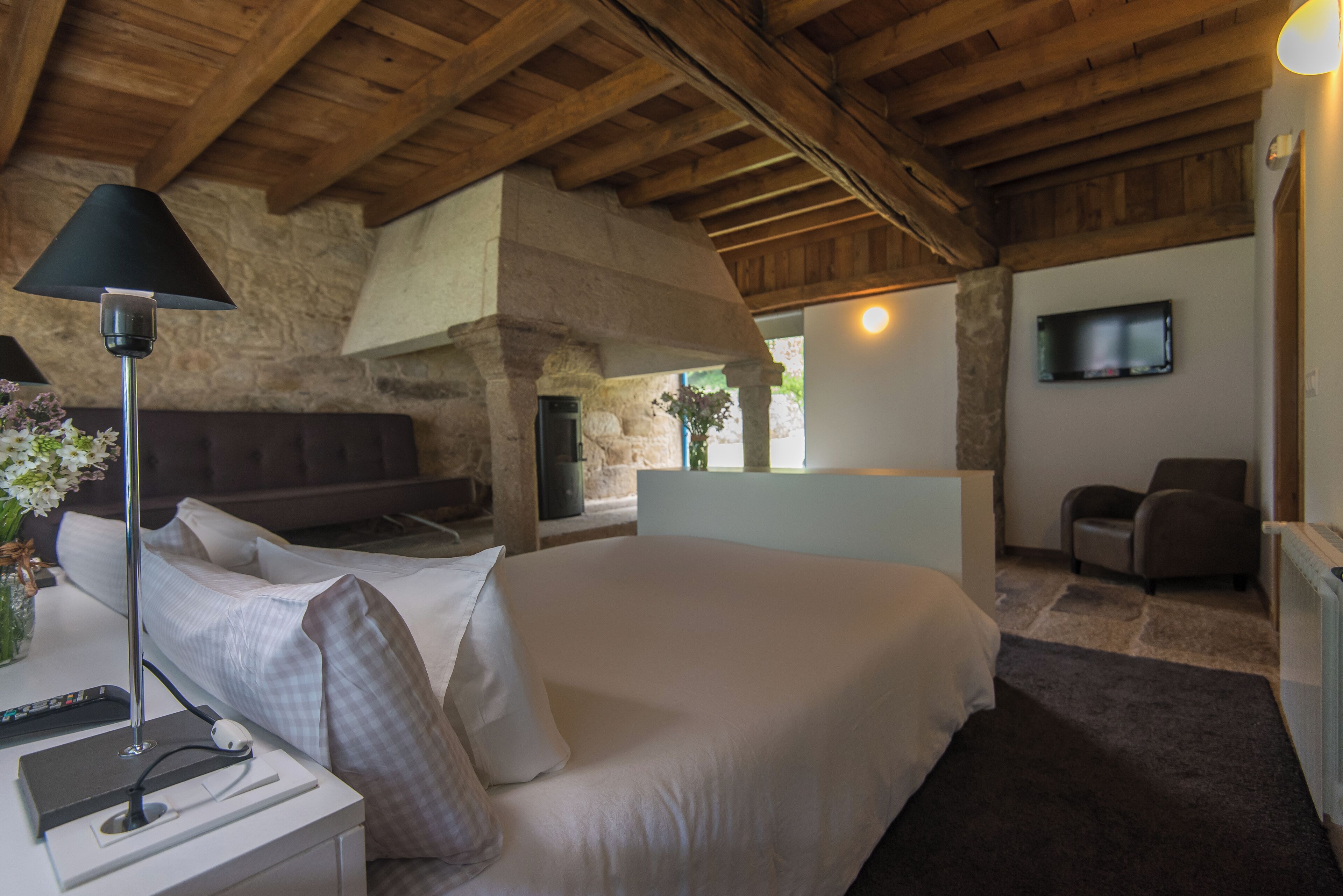 A bedroom with a large bed, a bedside table with a lamp, a TV mounted on the wall, and a stone fireplace.
