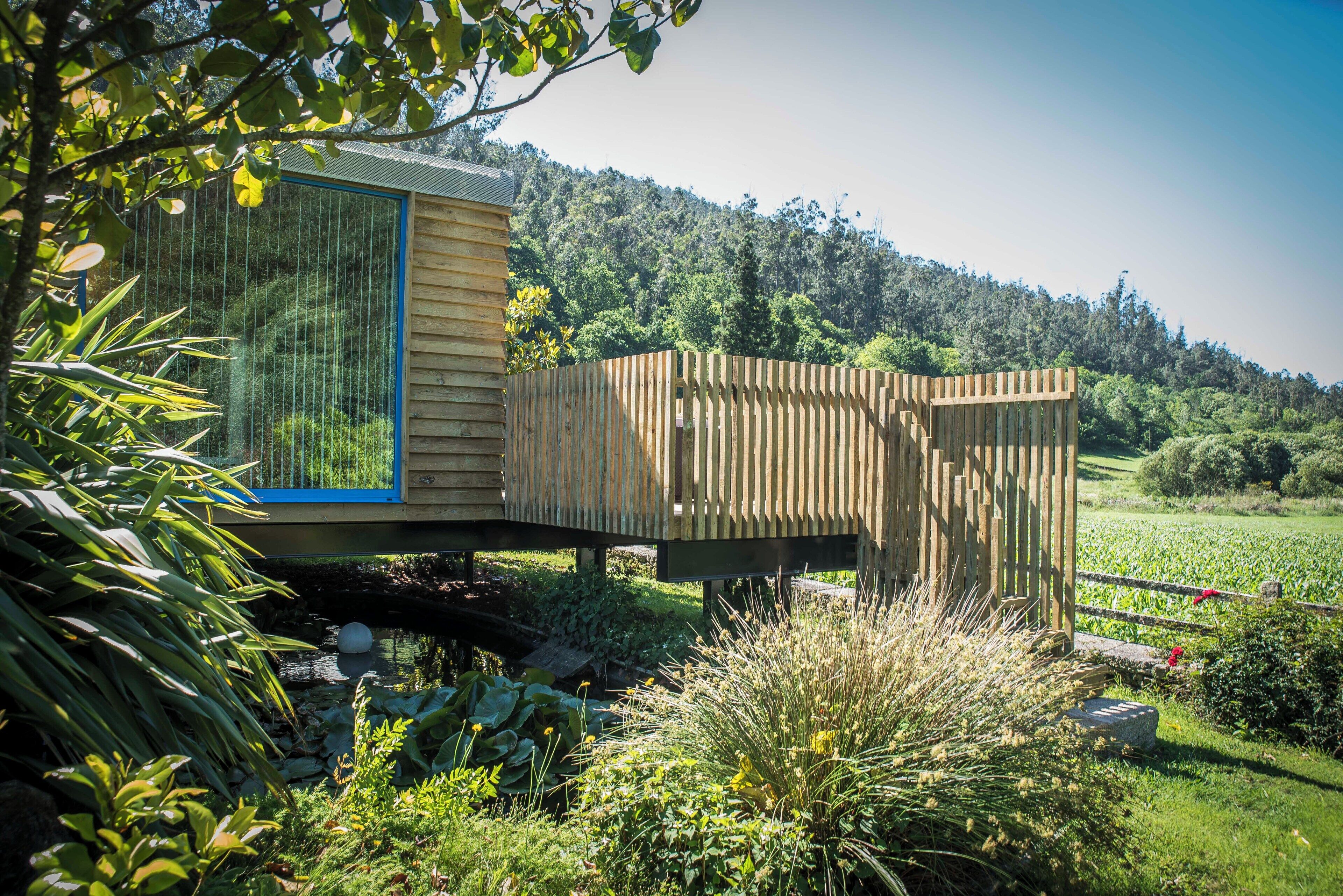 A wooden cabin with a glass door, surrounded by lush greenery and a hill in the background.