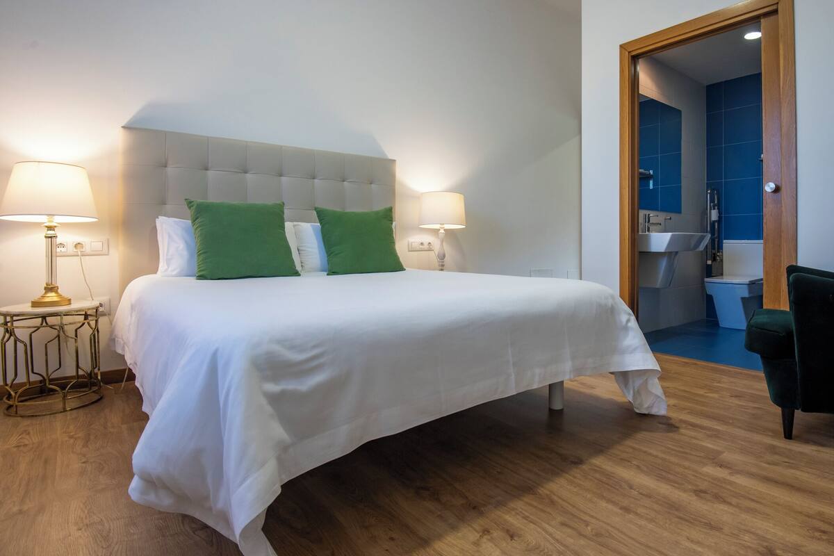 Une chambre d’hôtel avec un grand lit, deux lampes de chevet, un fauteuil vert et une salle de bain visible à travers une porte ouverte.