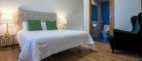 A hotel room with a large bed, two bedside lamps, a green chair, and a bathroom visible through an open door.