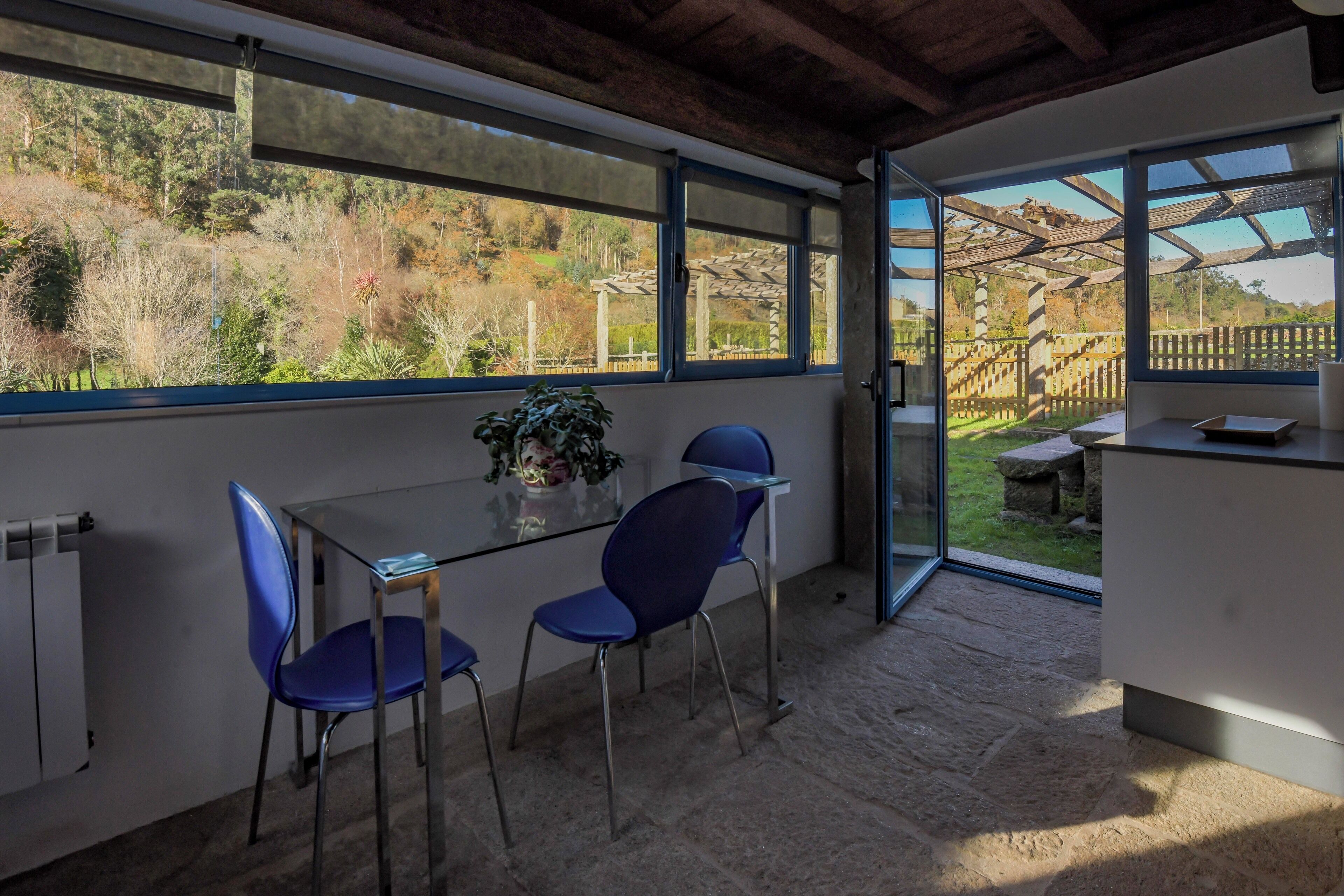 Una stanza con un tavolo e sedie in vetro, una pianta in vaso e vista su un giardino tramite ampie finestre.