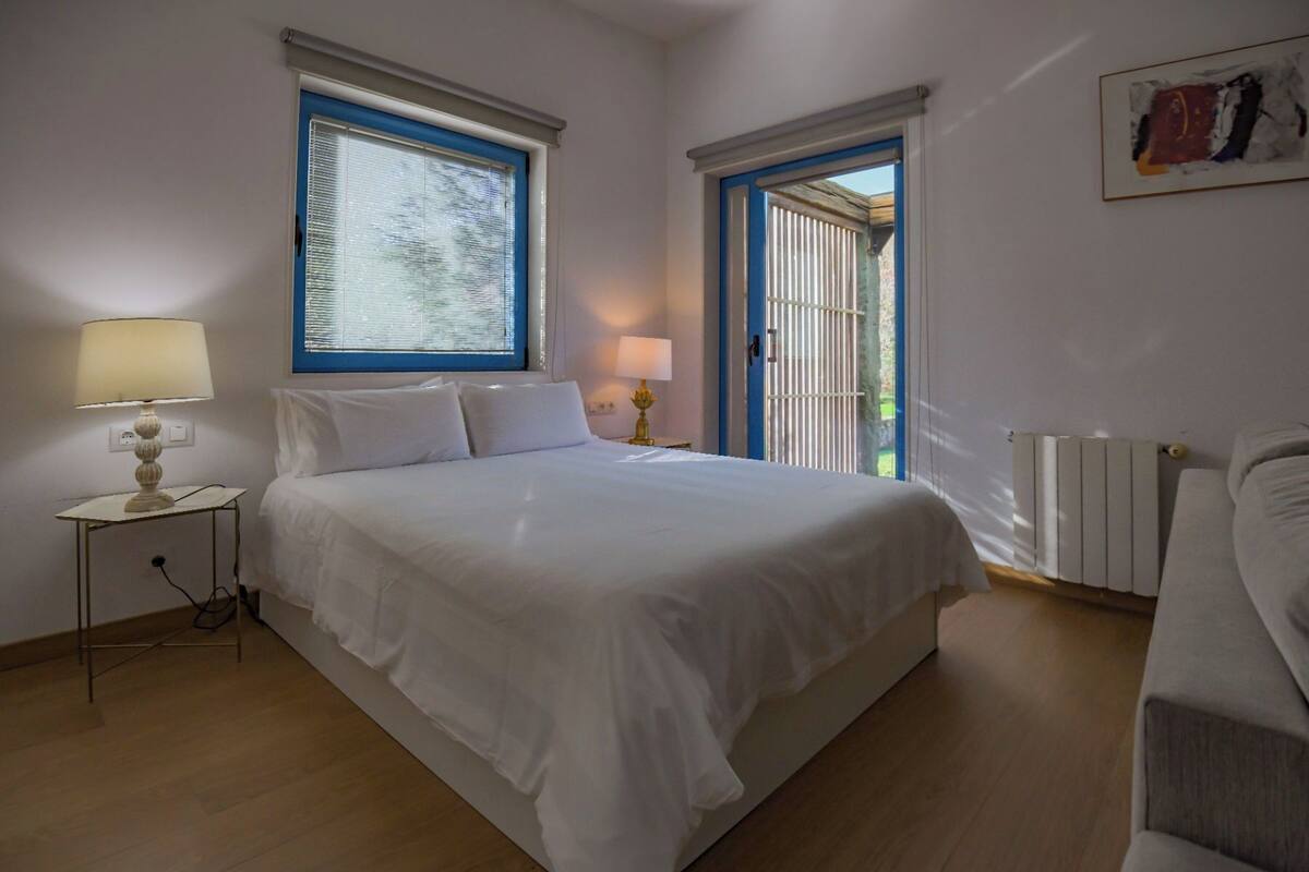 Une chambre à coucher comprenant un lit, deux tables de chevet avec des lampes, un radiateur et une porte donnant sur un espace extérieur.