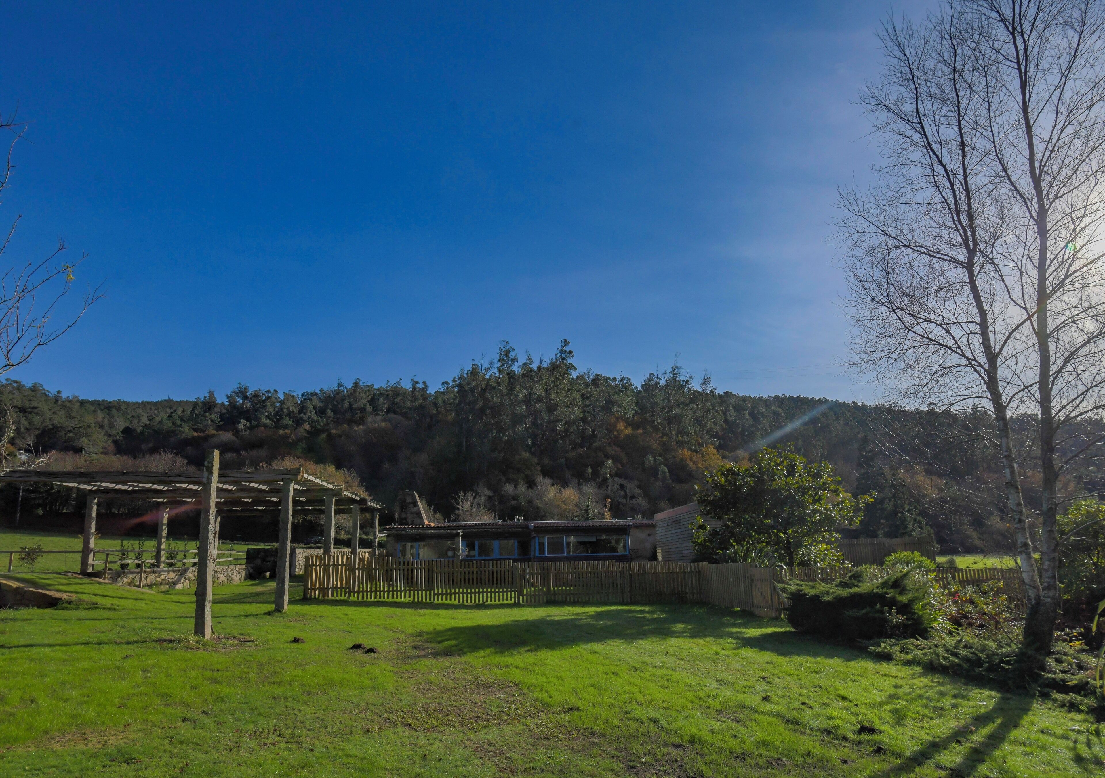 Una casa con terraza de madera, rodeada por un patio cercado y un cerro cubierto de bosque.