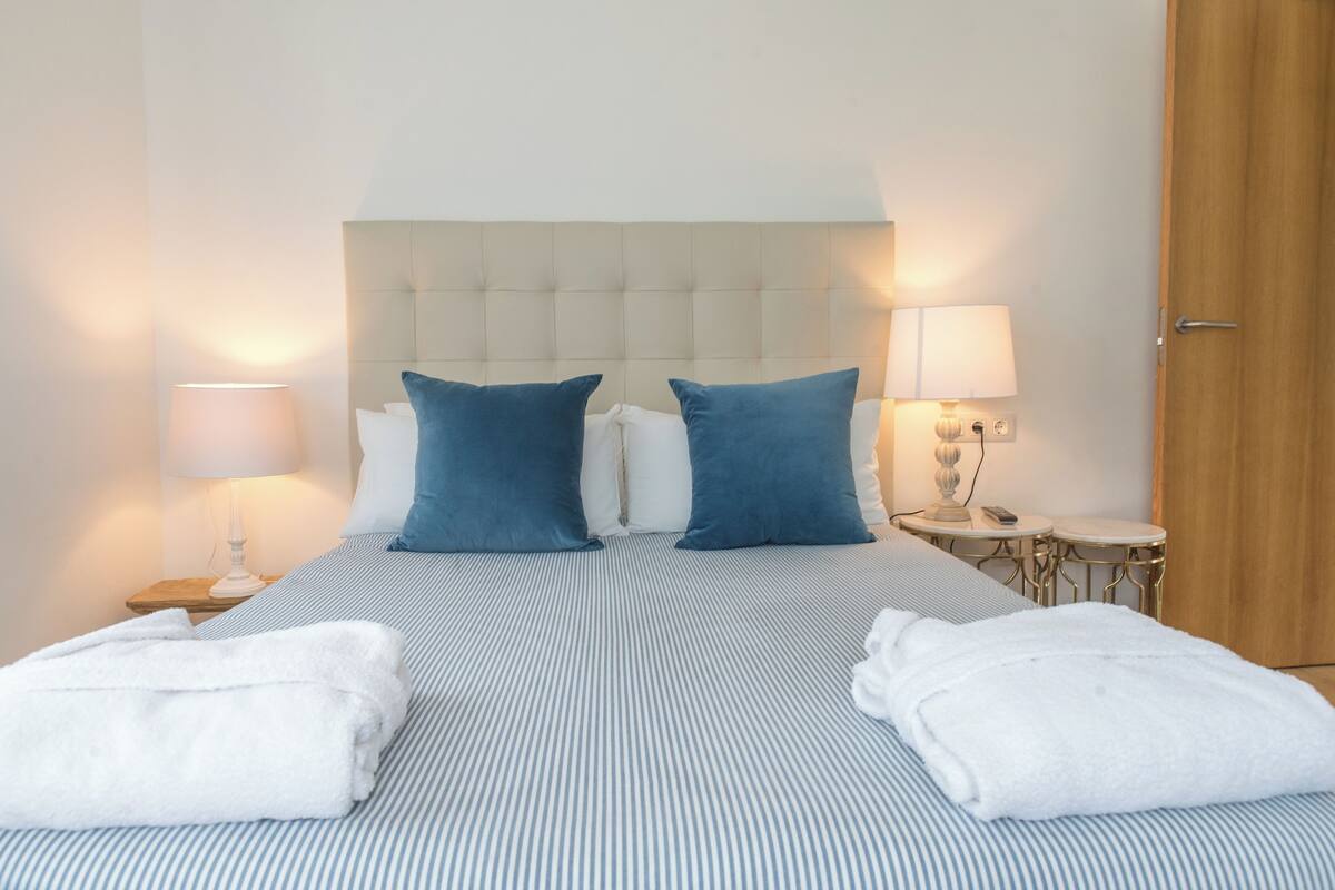 Une chambre d’hôtel avec un lit, deux oreillers, une table de chevet avec une lampe, une petite table avec une lampe et une porte en bois.