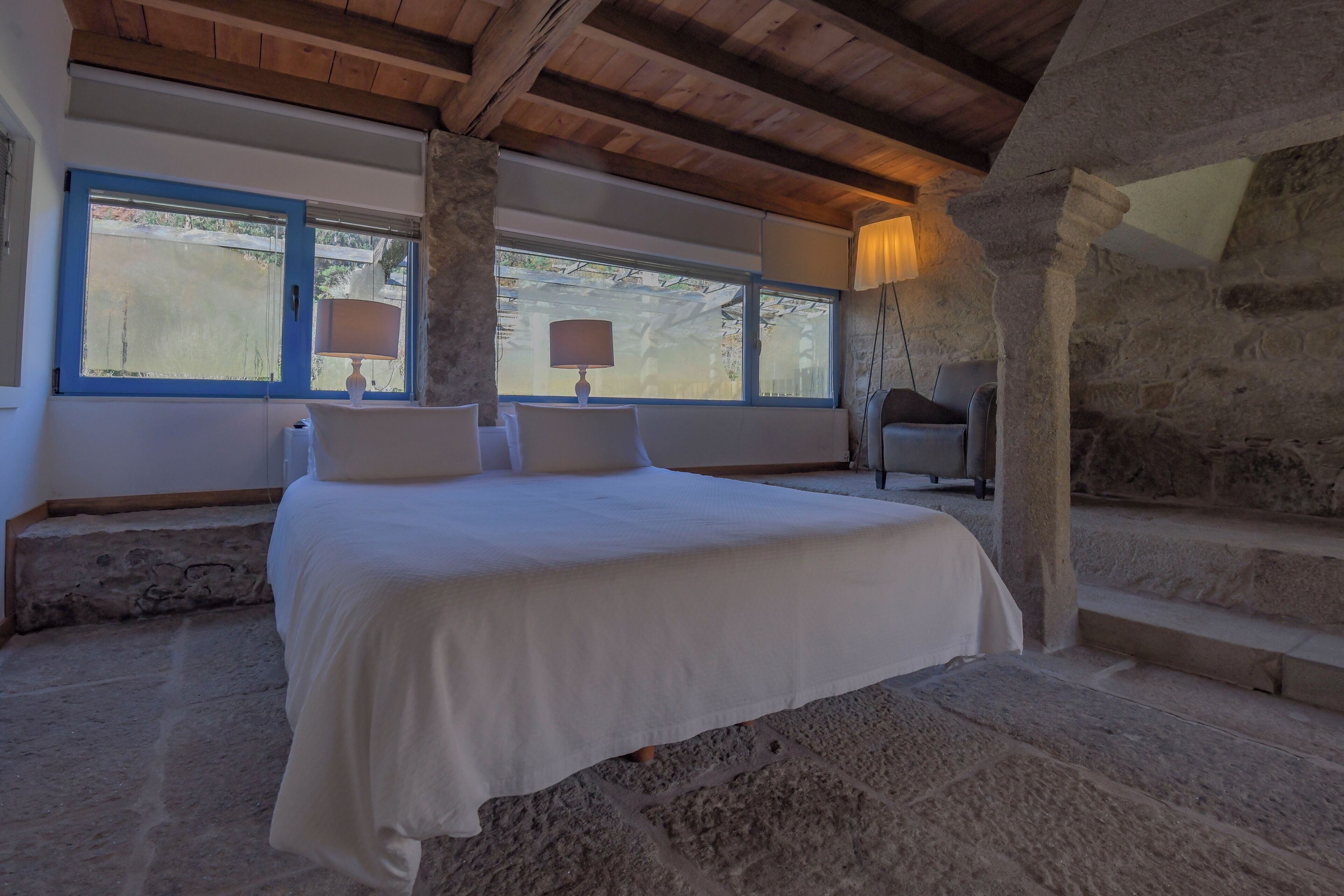 A bedroom with a stone floor, a bed with white linens, two windows with blue frames, and a stone pillar.