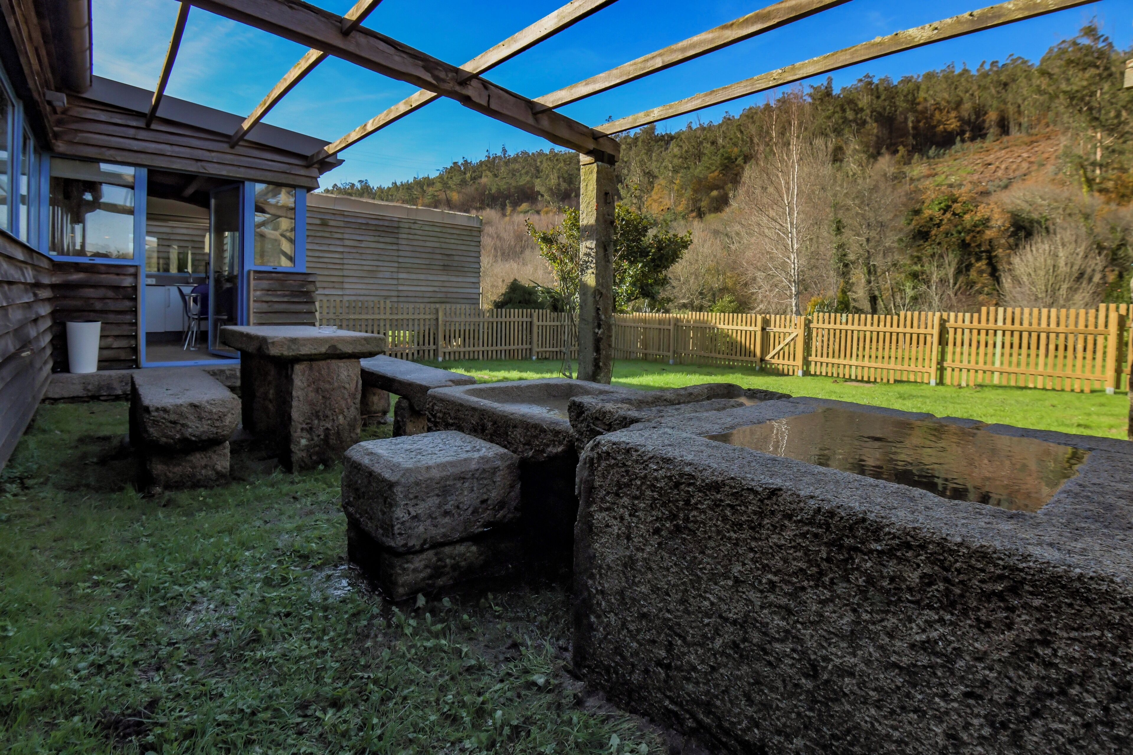 A wooden pergola with a stone bench and a small pond, surrounded by a wooden fence and a grassy area.