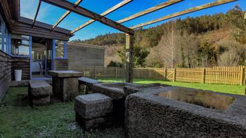 Una pérgola de madera con un banco de piedra y un pequeño estanque, rodeada por una valla de madera y una zona verde.