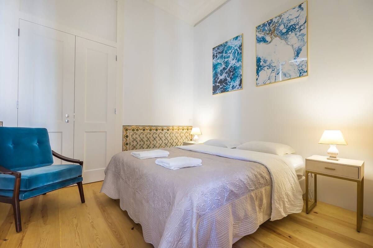 A neatly made bed with a white comforter and pillows, a blue armchair, a bedside table with a lamp, and two abstract paintings on the wall.