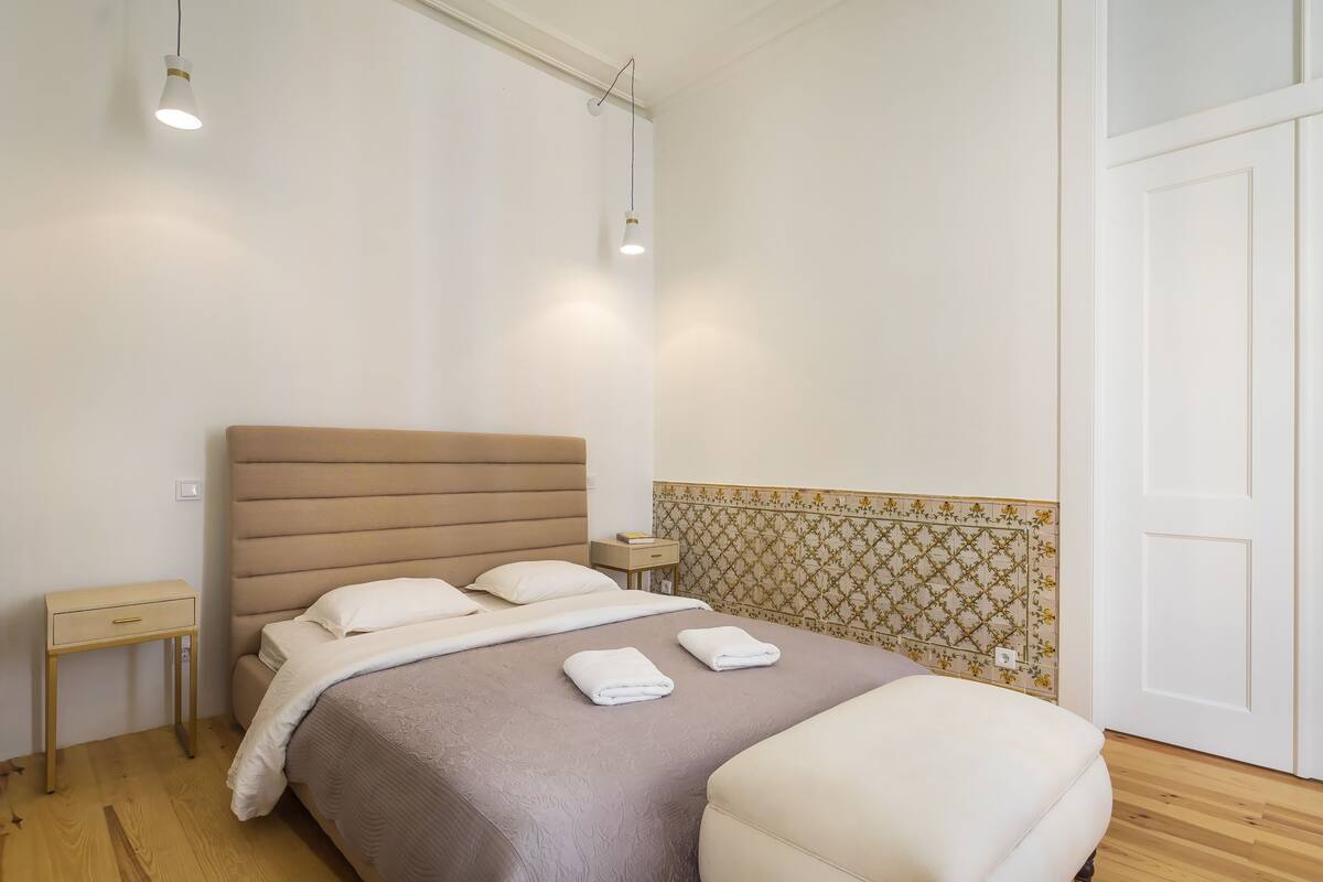 A neatly made bed with a headboard, bedside tables, and a patterned wall.