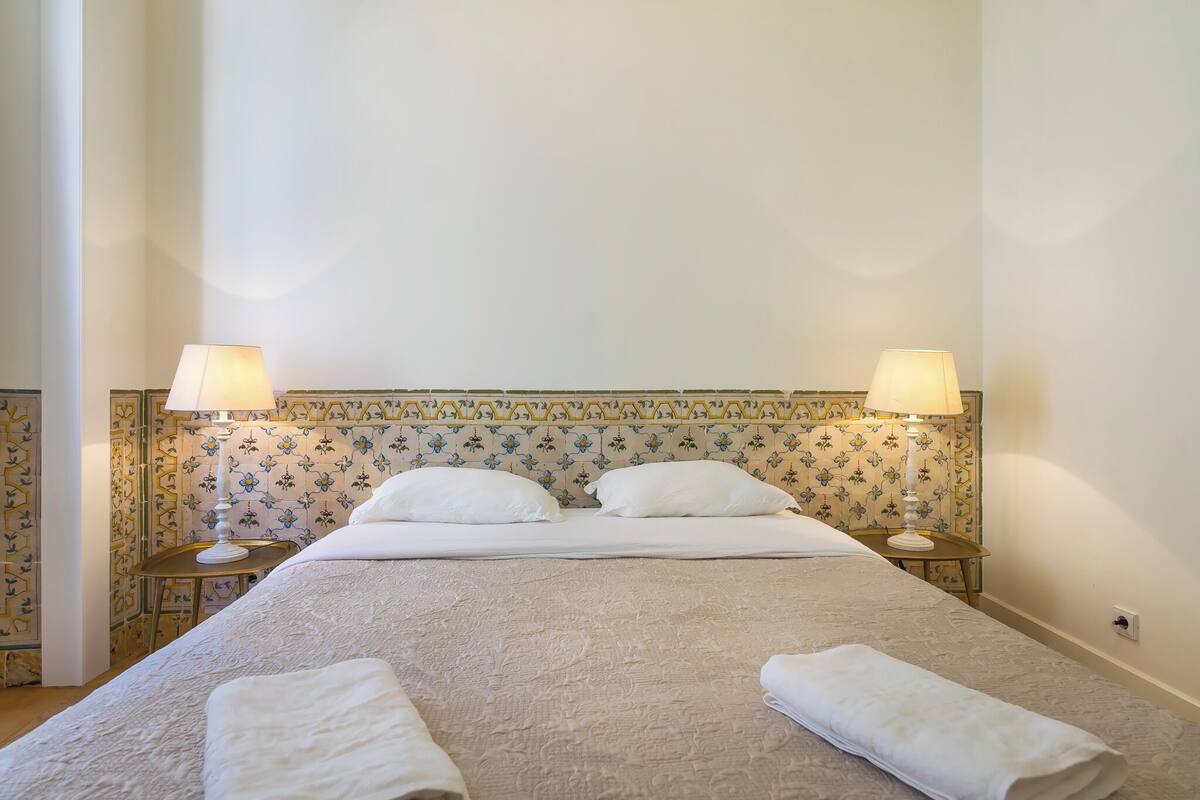 A neatly made bed with white linens and two bedside lamps, a floral-patterned headboard, and a small bedside table with a lamp.