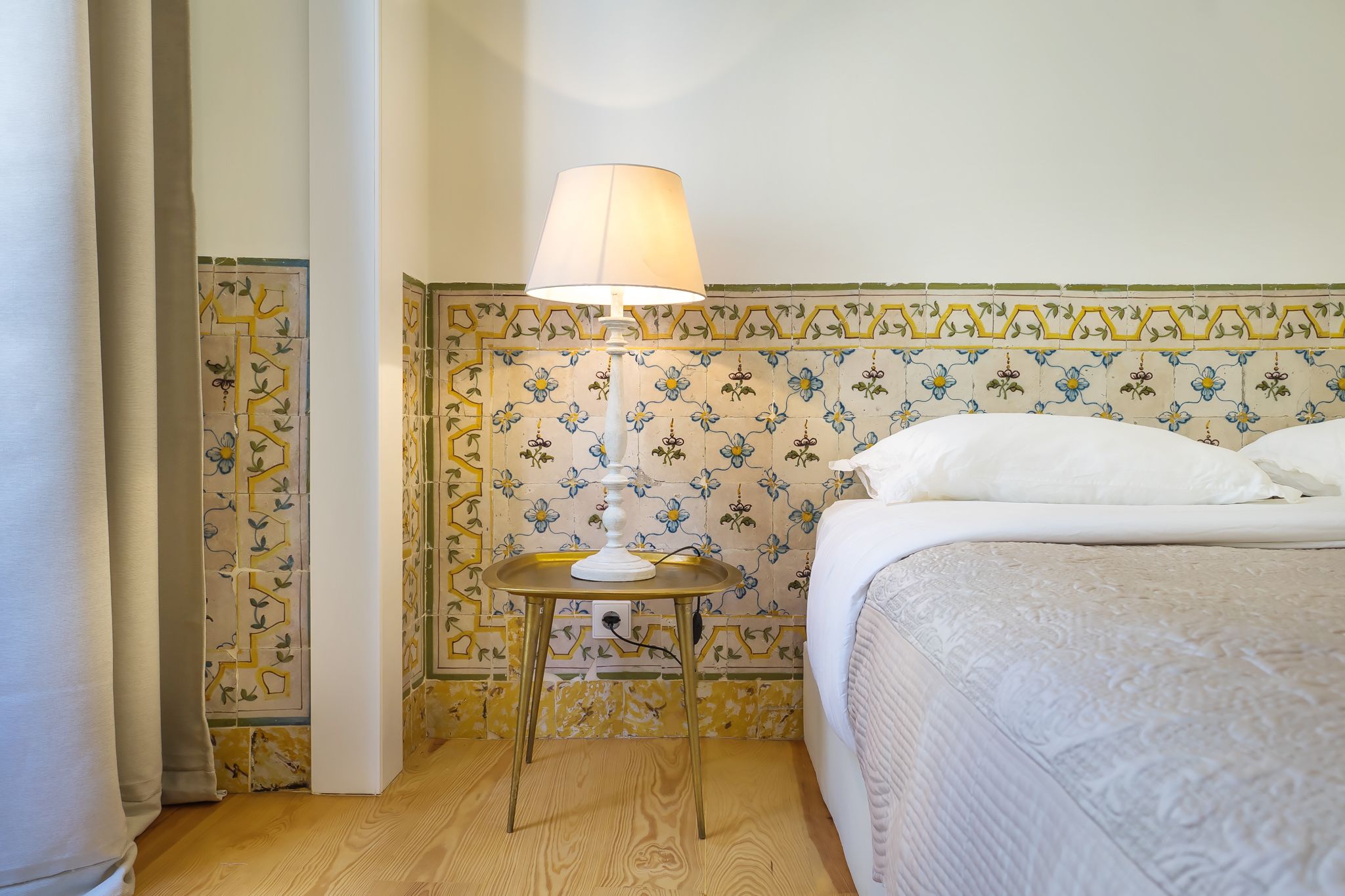 Una cama bien hecha con una colcha blanca y almohadas, una mesita de noche con una lámpara, y una pared con baldosas de estampado floral.