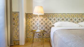 Una cama bien hecha con una colcha blanca y almohadas, una mesita de noche con una lámpara, y una pared con baldosas de estampado floral.