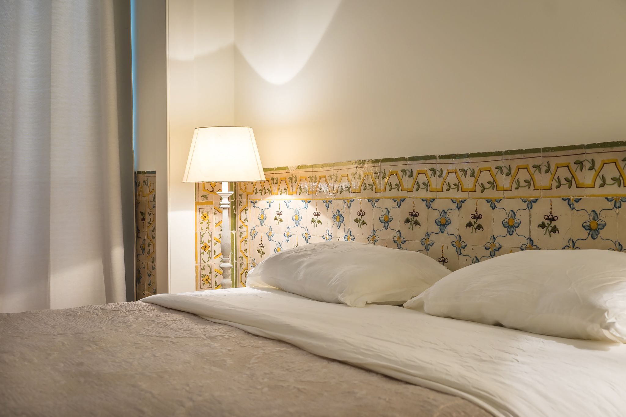 A neatly made bed with white linens and pillows, a bedside lamp, and a wall with a floral tile pattern.