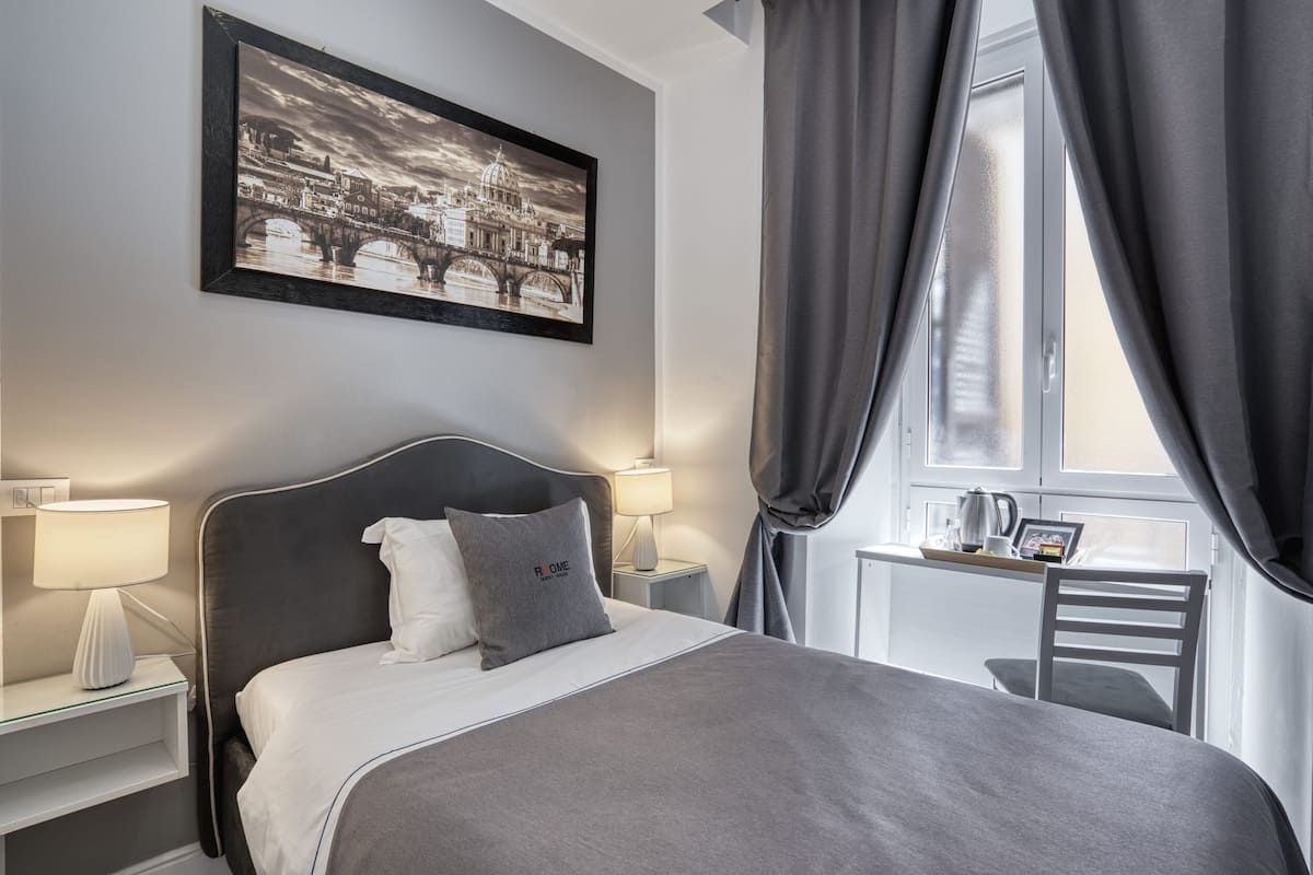 A neatly arranged bedroom with a bed, bedside lamps, a framed picture on the wall, and a window with curtains.
