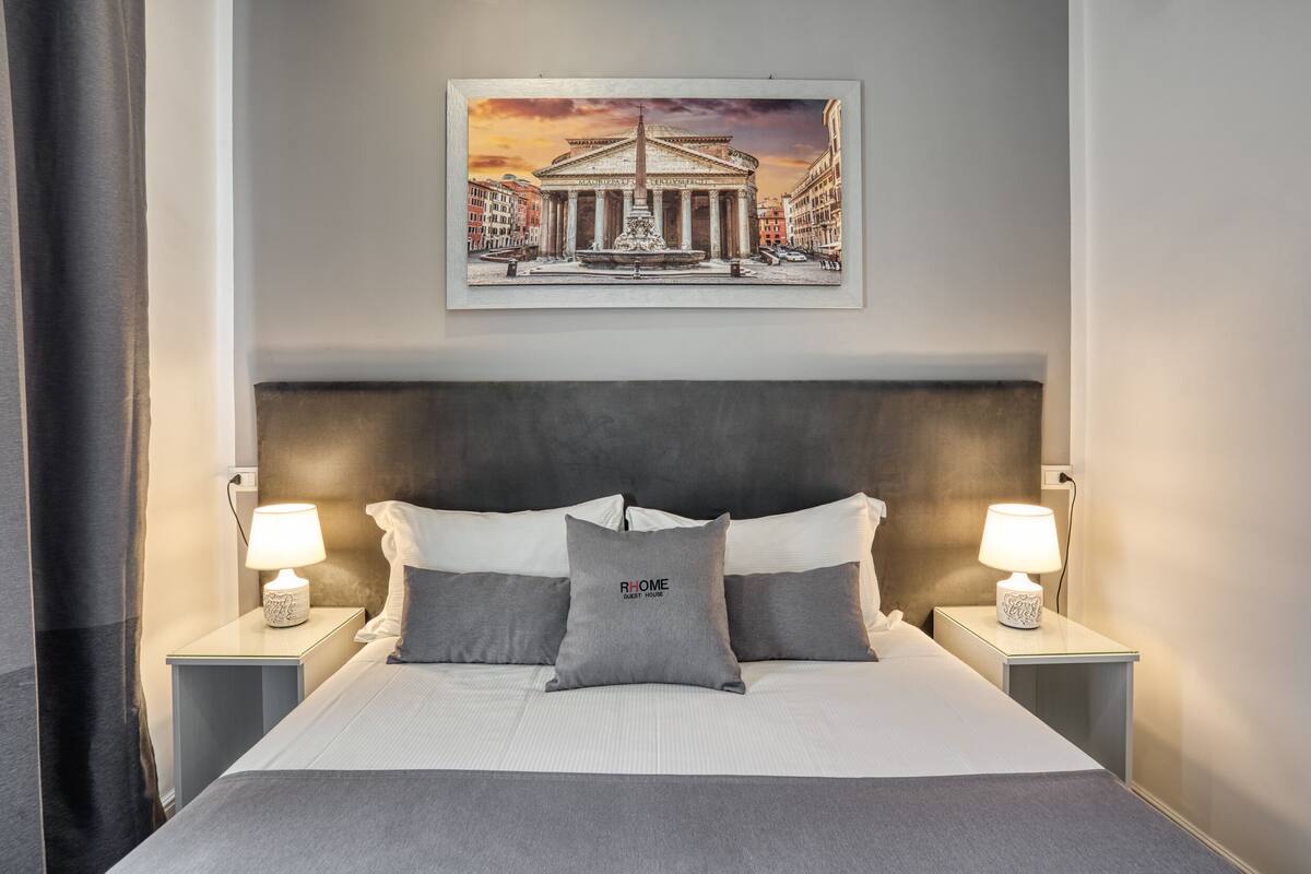 A hotel room with a bed, two bedside lamps, a painting of a classical building, and a grey pillow with "RHOME GUEST HOUSE" text.
