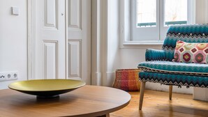 A room with a round wooden table, a colorful woven basket, a blue patterned sofa, and a white door. - The Color Hostel (Lisbon)