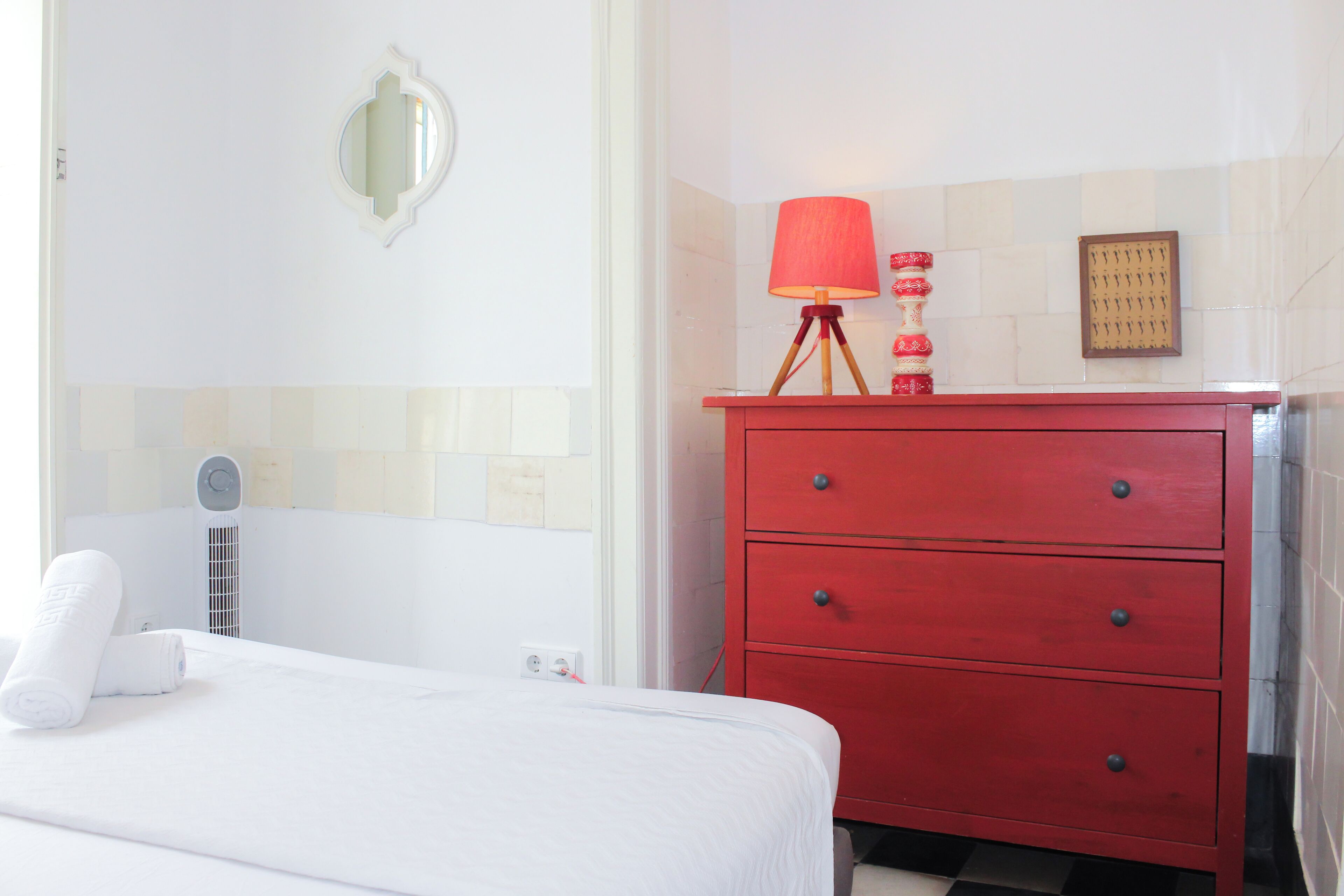 Une chambre d’hôtel avec un lit, une commode rouge, un miroir fixé au mur et une lampe rouge.