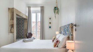 A hotel room with a bed, a chair, a nightstand, a lamp, a window with curtains, and a view of buildings outside. - The Color Hostel (Lisbon)