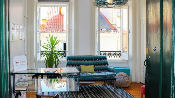 A room with a glass table, a blue patterned sofa, a striped rug, and a potted plant. - The Color Hostel (Lisbon)