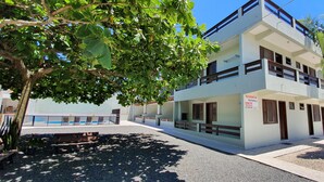 Un immeuble résidentiel à deux étages, doté de balcons, d’une pancarte indiquant qu’il est à vendre, et d’une zone pavée avec des bancs et des arbres.