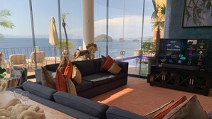 A modern living room with a sofa, cushions, a coffee table, and a TV stand with a screen displaying various channels. - Luxurious Oceanfront Estate on Vallarta's South Shore, Pool, Jacuzzi, Chef, Maid (Puerto Vallarta)