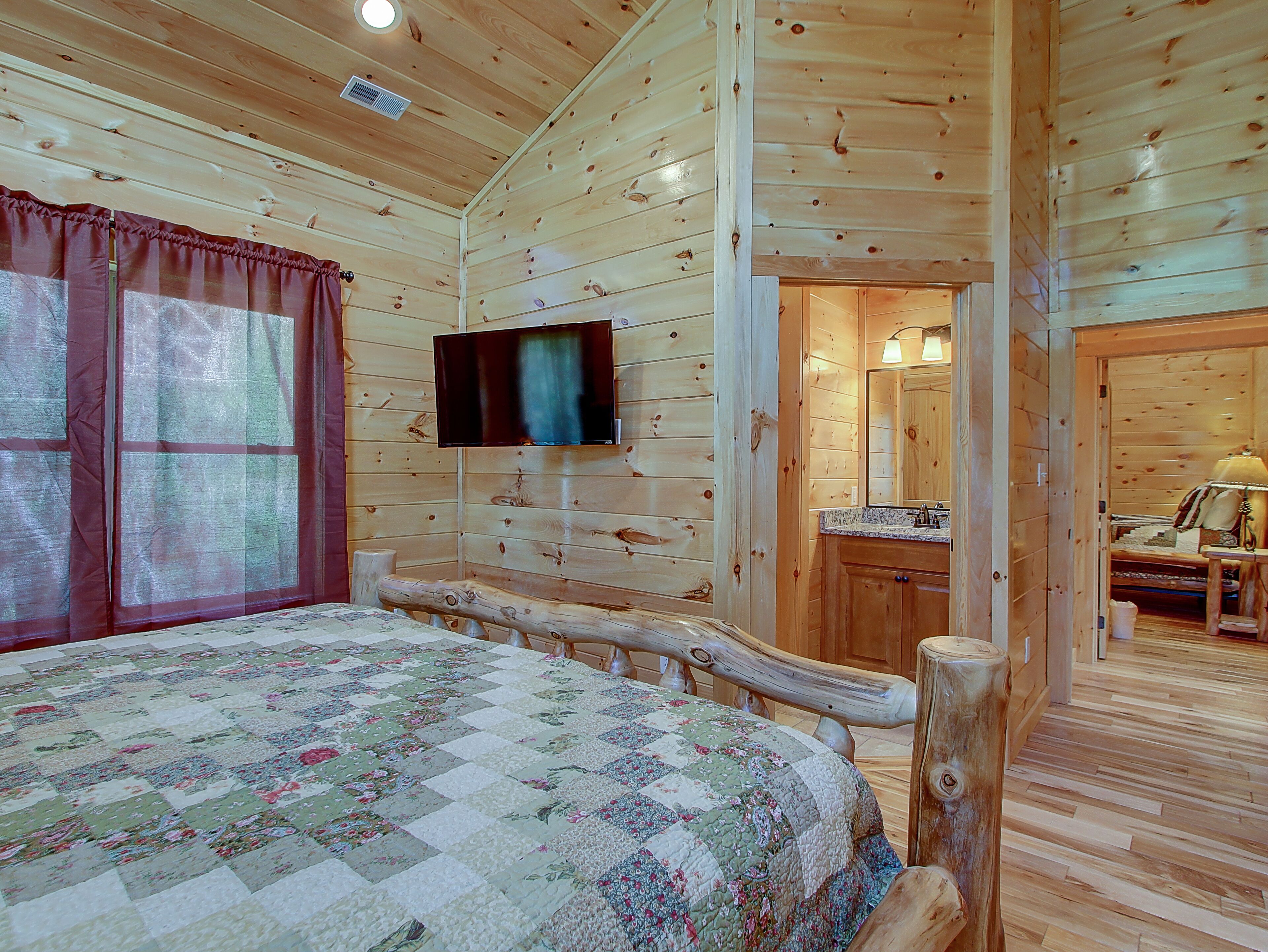 A wooden cabin bedroom with a bed, a television mounted on the wall, a bathroom with a sink and mirror, and a wooden bench.