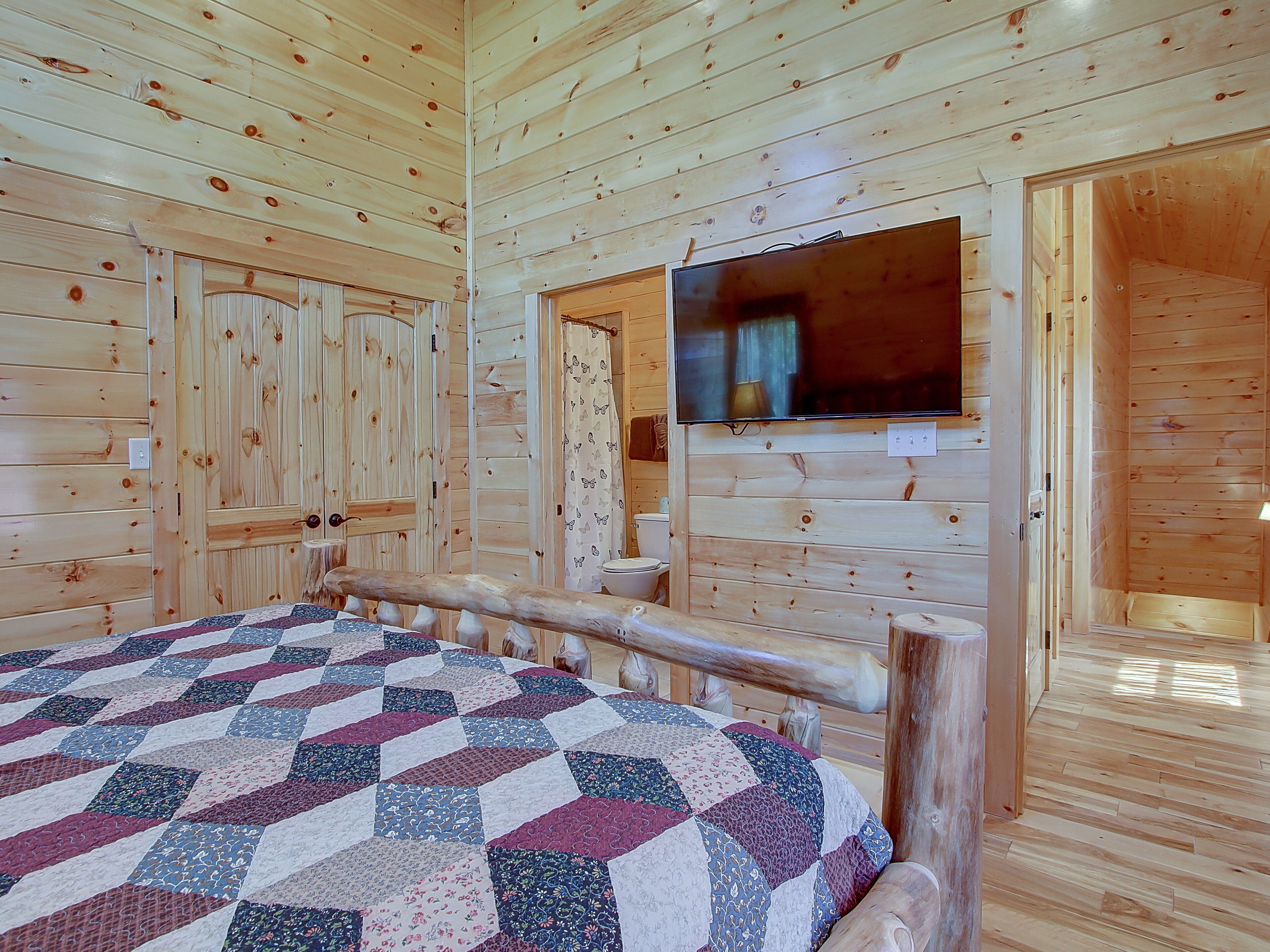A wooden cabin room with a bed, a television mounted on the wall, and a wooden door.