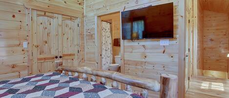 A wooden cabin room with a bed, a television mounted on the wall, and a wooden door.