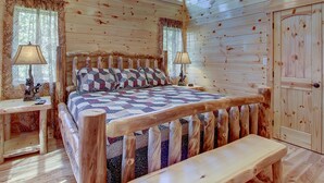 A log cabin bedroom with a wooden bed, two bedside lamps, a bench, and a window with curtains. - Mountain view, sleeps 14, pool table, arcade game, and hot tub! Plenty of space! (Gatlinburg)