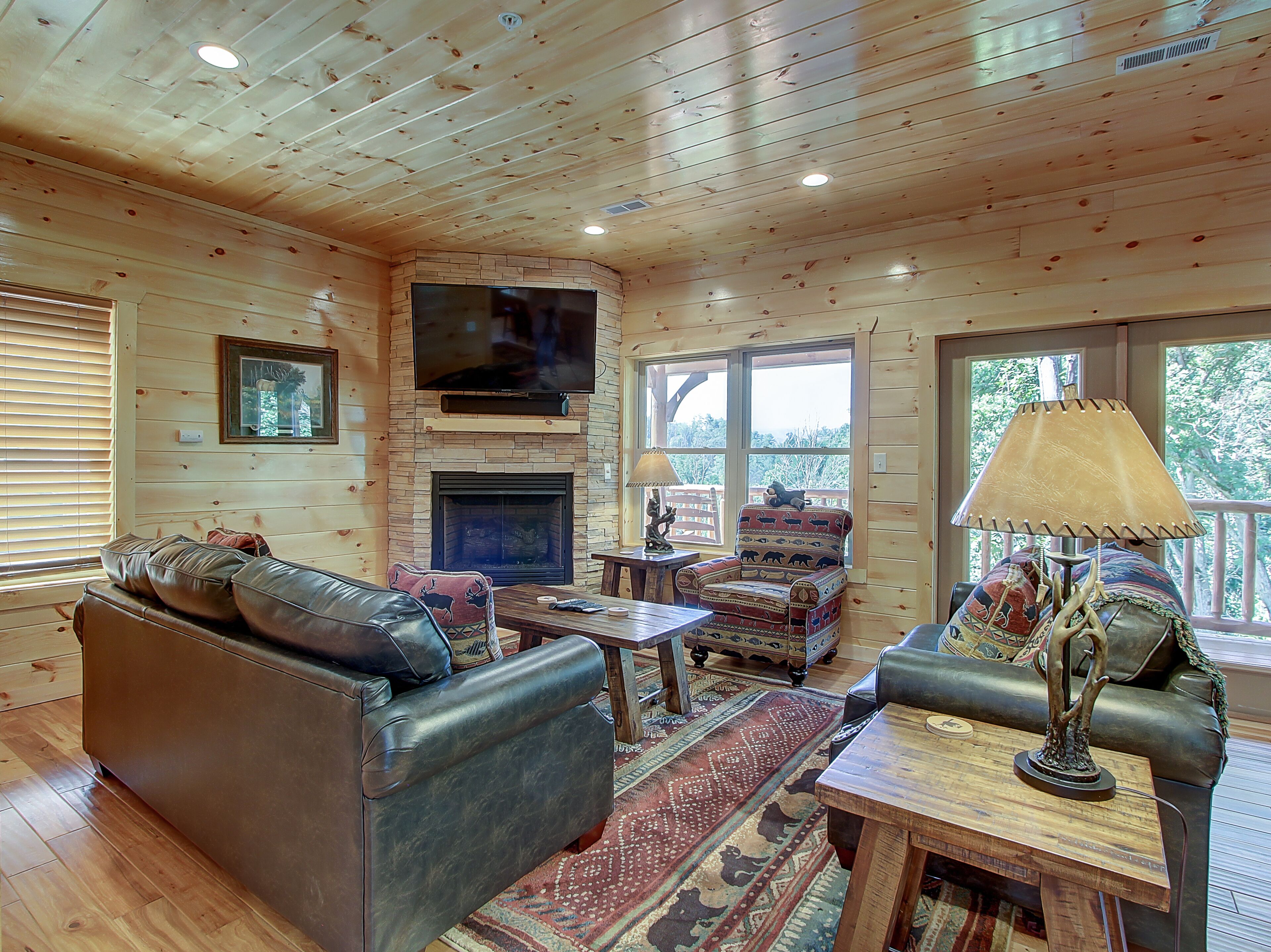 A cozy cabin interior with a leather sofa, a fireplace, a television mounted above it, and a large window offering a view of the outdoors.