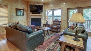 A cozy cabin interior with a leather sofa, a fireplace, a television mounted above it, and a large window offering a view of the outdoors. - Mountain view, sleeps 14, pool table, arcade game, and hot tub! Plenty of space! (Gatlinburg)