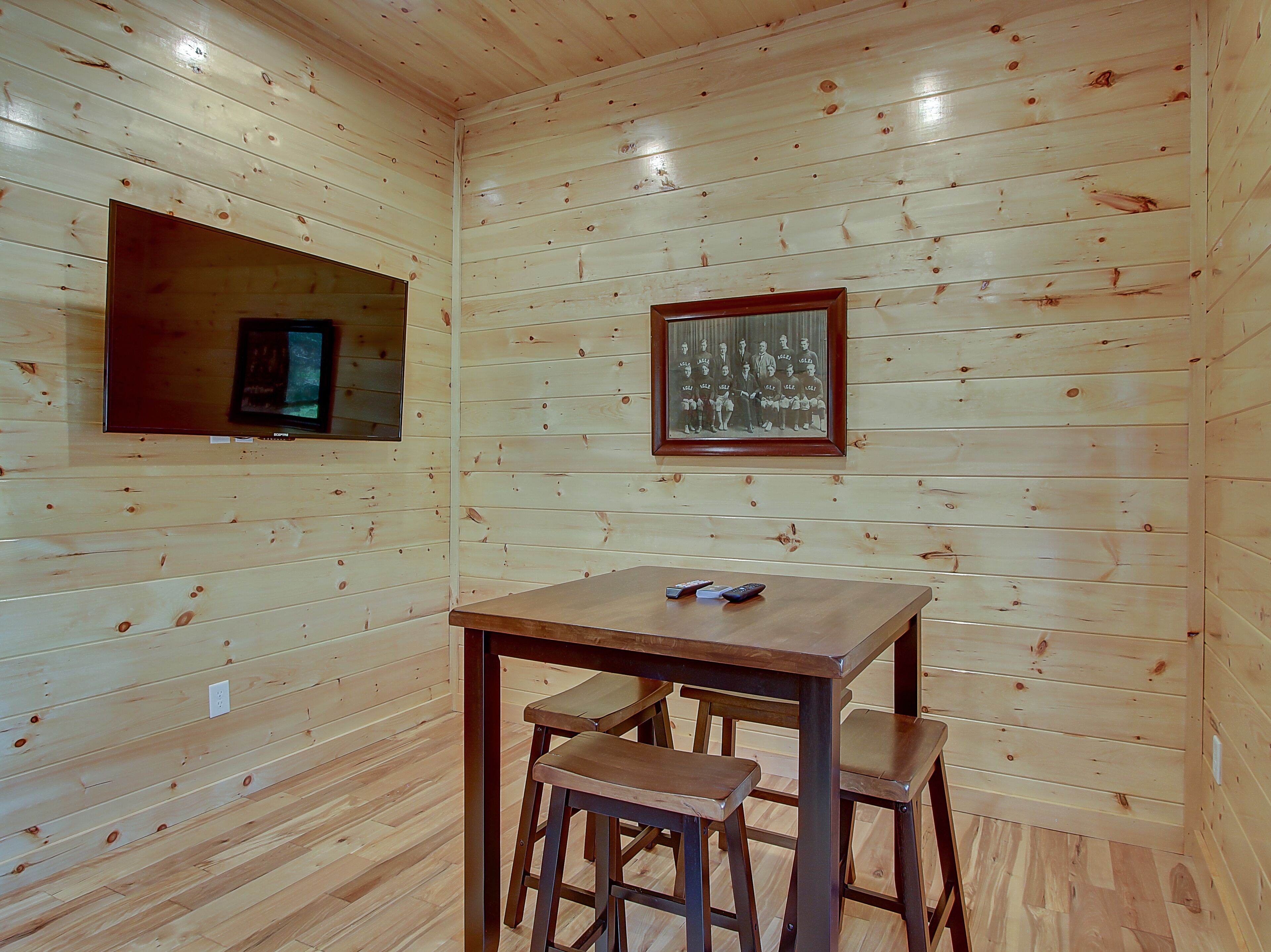 A wooden-paneled room with a table and two chairs, a framed photograph on the wall, and a television mounted on the wall.