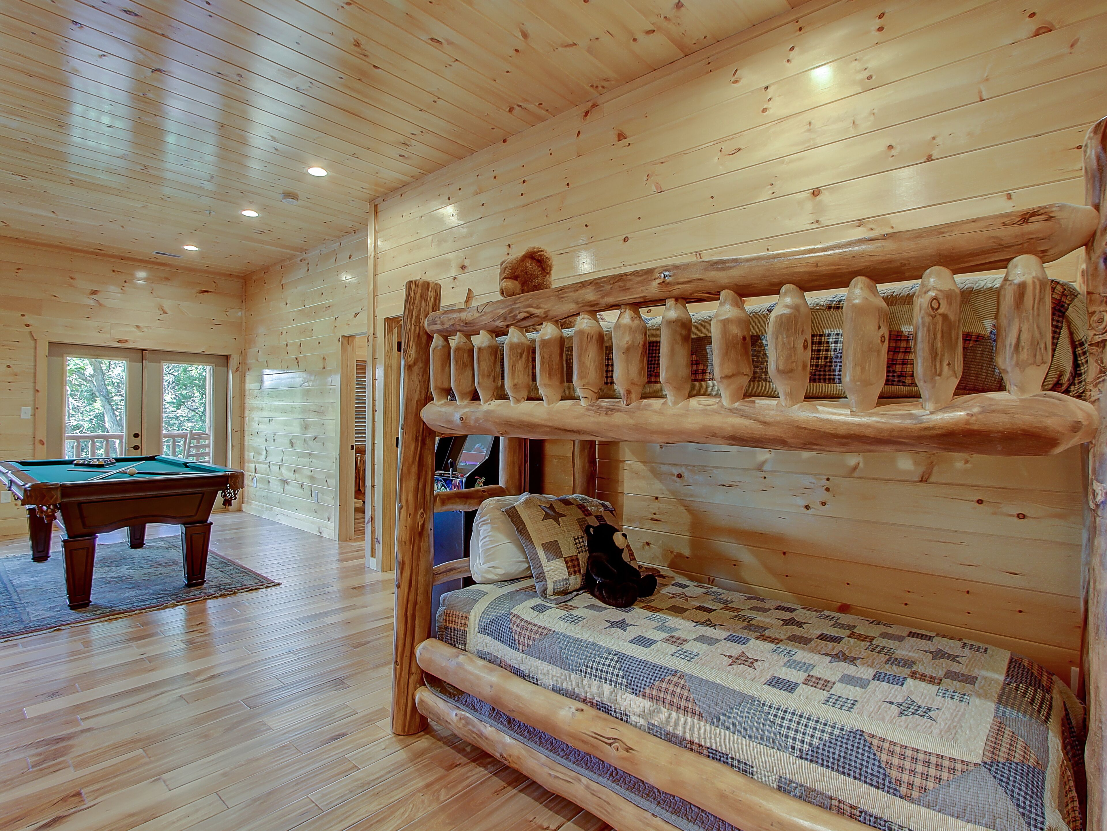 A wooden cabin interior with a pool table, a bunk bed with a quilt, and a door leading outside.