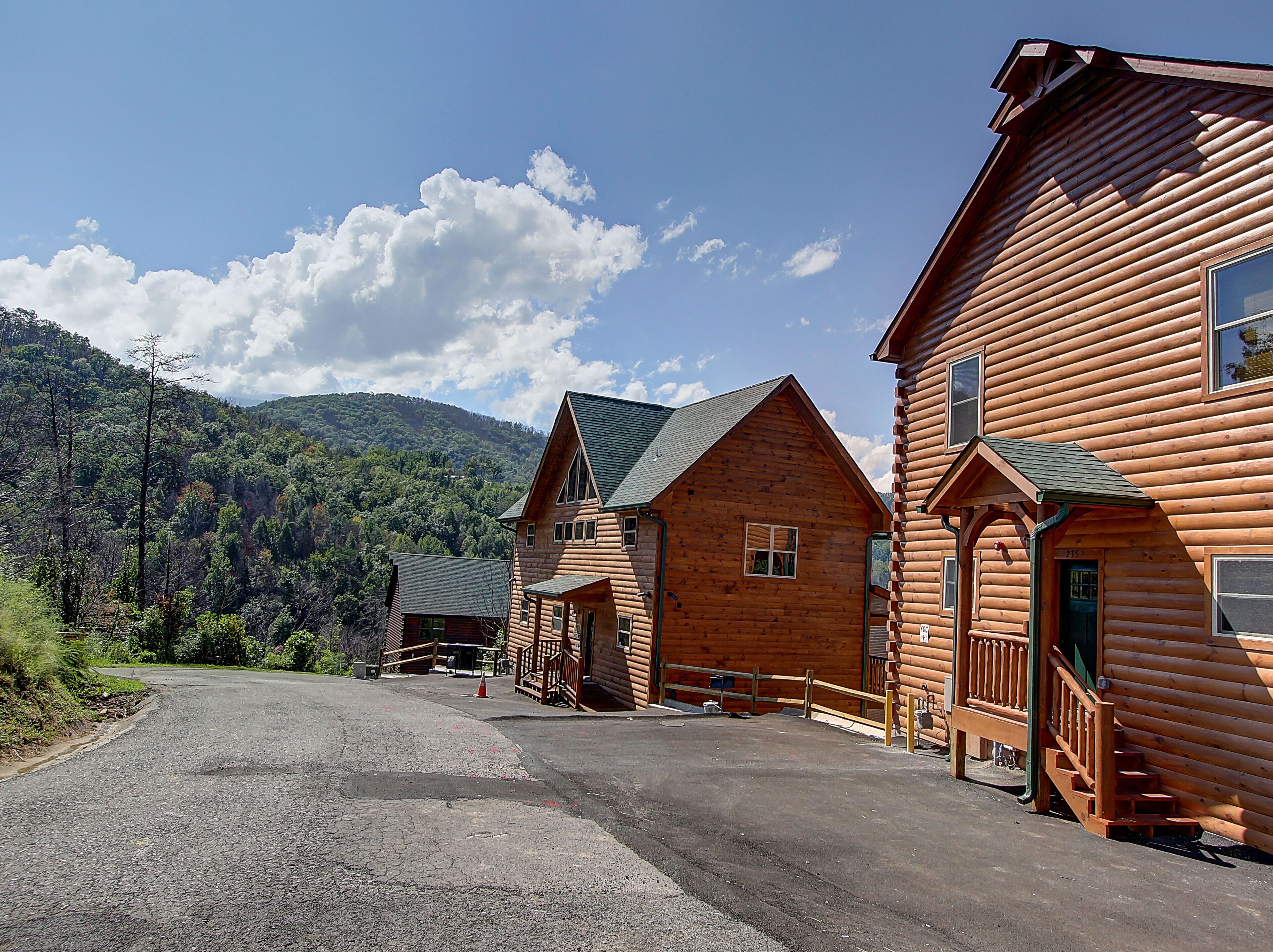 A log cabin with a porch and a garage, surrounded by a gravel driveway and forested hills.