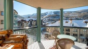A balcony with a view of a snowy mountain landscape, wooden logs, and a table with chairs. - Centennial 2 Bedroom Condo Unit no. 13ab (Avon)