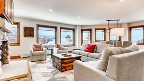 A spacious living room with a stone fireplace, large windows, and a view of a snowy landscape. - Centennial 2 Bedroom Condo Unit no. 13ab (Avon)