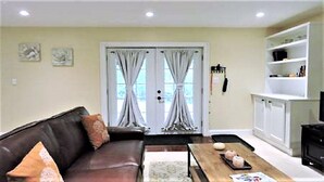 A living room with a brown leather sofa, a wooden coffee table, and a television on a cabinet. There are curtains on the windows and a shelf with items on the wall.